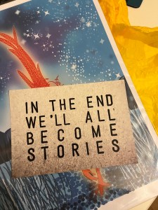 postcard with text only that reads in the end we all become stories, post card is in the centre of the image obscuring most of an artist print of a pheasant (colour is luminous oranges and reds) with starry sky
