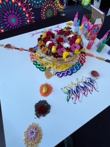 background purple with colourful shapes and patterns, a table in the foreground has a bowl with flowers beautifully displayed surrounded by patterns in coloured sand, flowers, flower petals and lights 